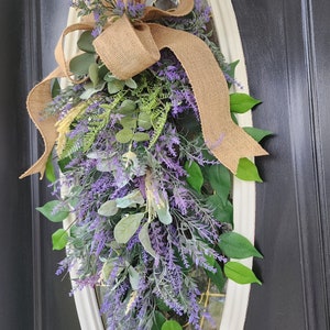 Peut inclure: Une couronne d&eacute;corative en forme de goutte d'eau avec de la lavande artificielle, des feuilles vertes et un n&oelig;ud en toile de jute. La couronne est fix&eacute;e &agrave; une fen&ecirc;tre ovale encadr&eacute;e de blanc sur une porte noire. La lavande est violette.