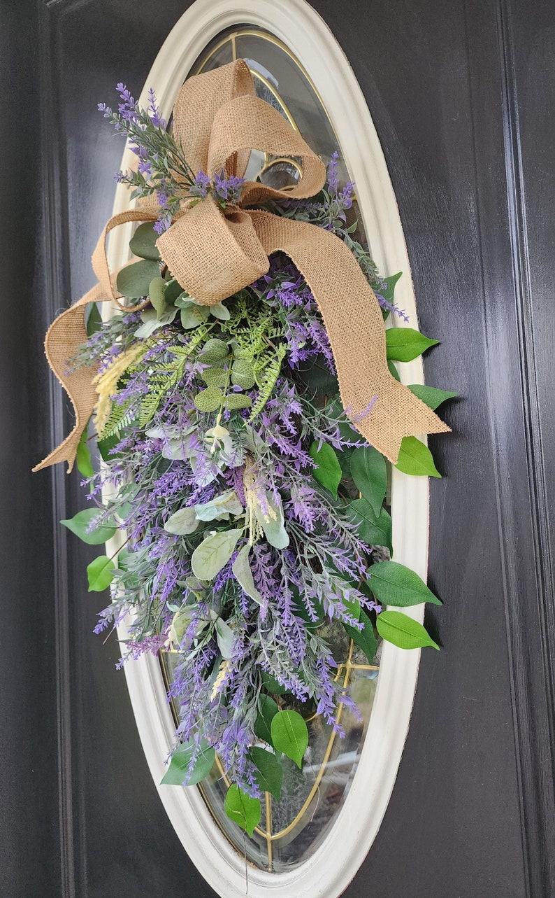 Peut inclure: Une couronne d&eacute;corative en forme de goutte d'eau avec de la lavande artificielle, des feuilles vertes et un n&oelig;ud en toile de jute. La couronne est fix&eacute;e &agrave; une fen&ecirc;tre ovale encadr&eacute;e de blanc sur une porte noire. La lavande est violette.