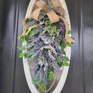 Peut inclure: Une couronne de porte ovale avec un cadre cr&egrave;me, orn&eacute;e de lavande violette, de feuilles vertes et d'un n&oelig;ud en toile de jute. L'arrangement floral est con&ccedil;u pour une porte et apporte une touche naturelle &agrave; l'entr&eacute;e d'une maison.