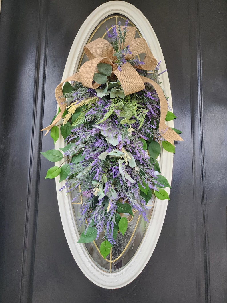 Peut inclure: Une couronne de porte ovale avec un cadre cr&egrave;me, orn&eacute;e de lavande violette, de feuilles vertes et d'un n&oelig;ud en toile de jute. L'arrangement floral est con&ccedil;u pour une porte et apporte une touche naturelle &agrave; l'entr&eacute;e d'une maison.