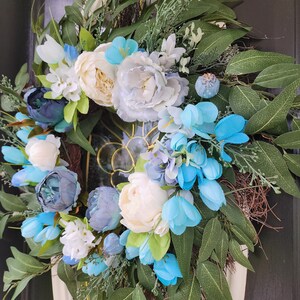 May include: A blue and white floral wreath with white peonies, blue hydrangeas, and green leaves. The wreath is hanging on a black door.