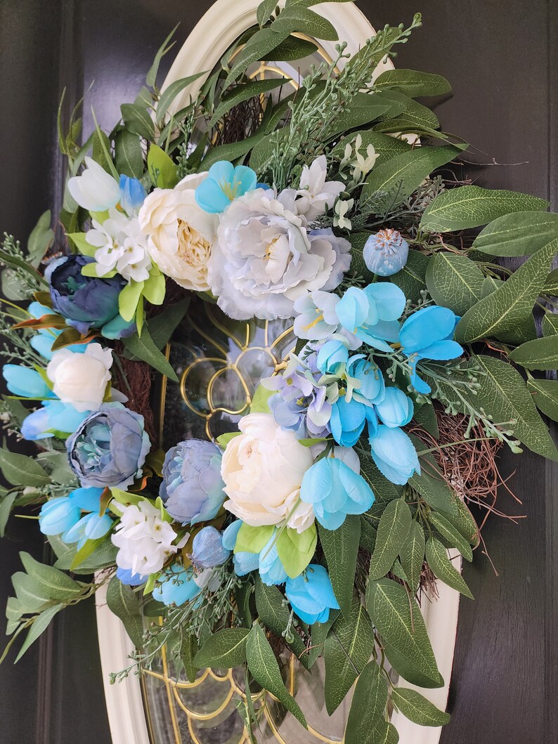 May include: A blue and white floral wreath with white peonies, blue hydrangeas, and green leaves. The wreath is hanging on a black door.