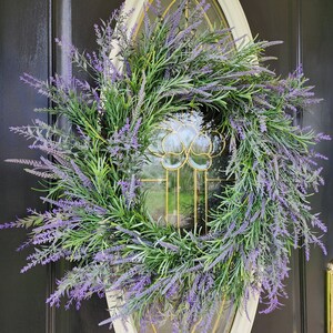 May include: A lavender wreath with purple flowers and green leaves hangs on a black door with a white oval frame and a glass window.