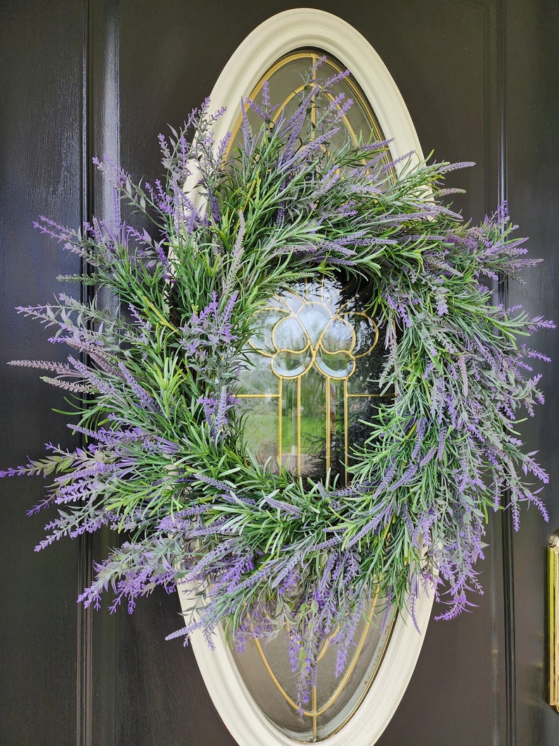 May include: A lavender wreath with purple flowers and green leaves hangs on a black door with a white oval frame and a glass window.