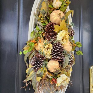 Op de afbeelding: Een herfst-deurkrans met nep-pompoenen, dennenappels en bladeren in tinten van goud, bruin en groen. De krans hangt aan een zwarte deur.
