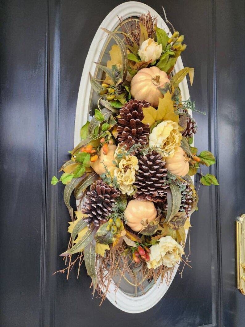 Op de afbeelding: Een herfst-deurkrans met nep-pompoenen, dennenappels en bladeren in tinten van goud, bruin en groen. De krans hangt aan een zwarte deur.