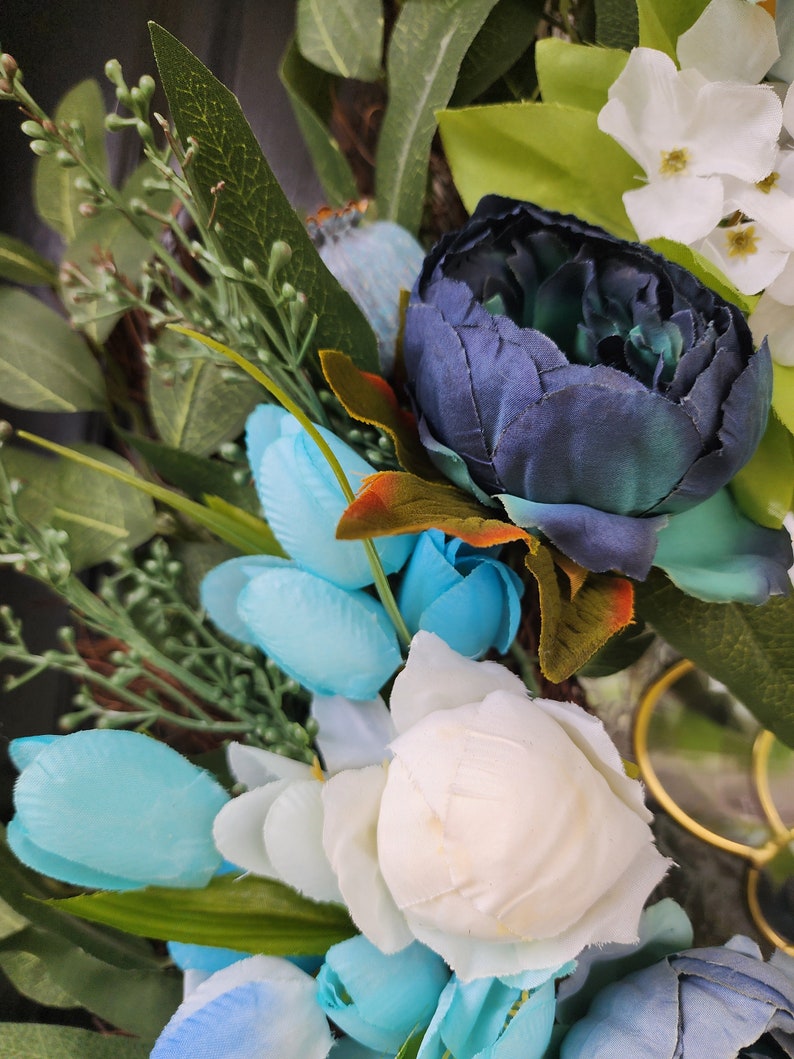 May include: A wreath featuring blue, white and green flowers. The wreath is made from artificial flowers and greenery.