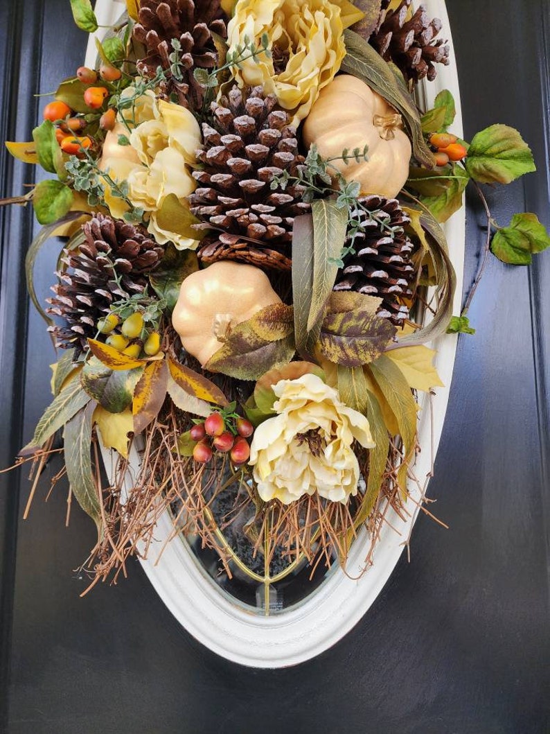 Op de afbeelding: Een herfst-deurkrans met nep-pompoenen, dennenappels en bloemen in tinten van bruin, goud en cr&egrave;me.