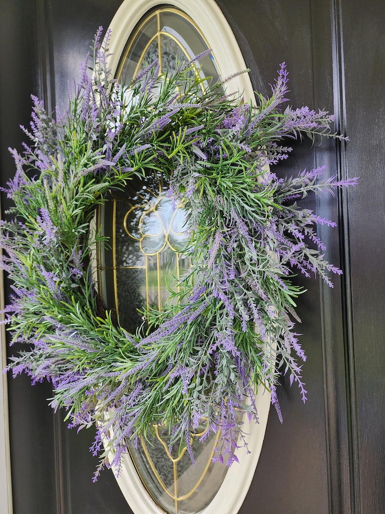 May include: A purple and green lavender wreath with a white oval frame. The wreath is hanging on a black door.