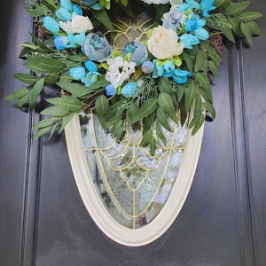 May include: A blue and white floral wreath with green leaves hangs on a black door. The wreath is made of artificial flowers and greenery.