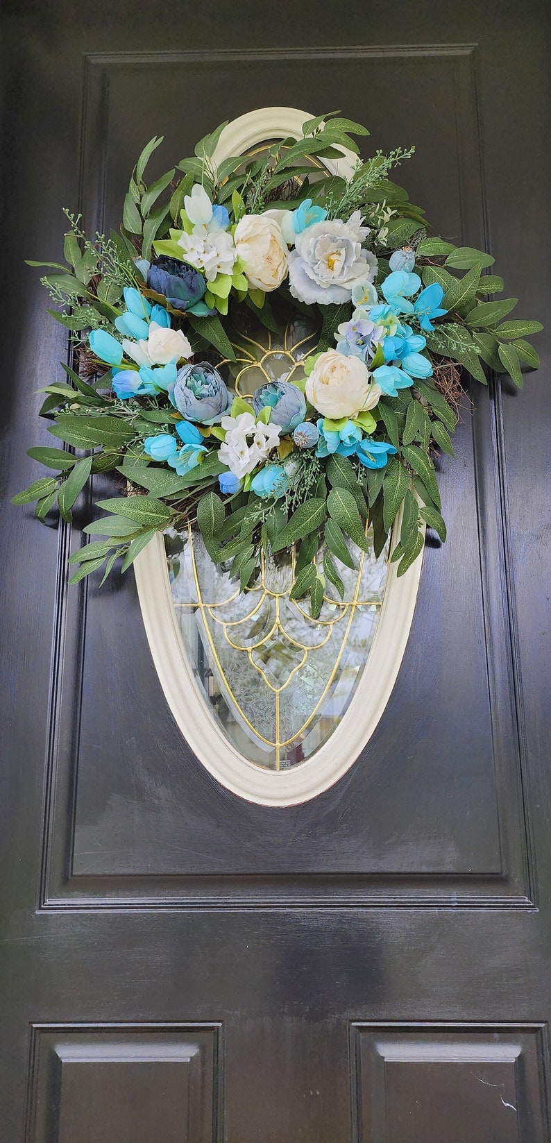 May include: A blue and white floral wreath with green leaves hangs on a black door. The wreath is made of artificial flowers and greenery.