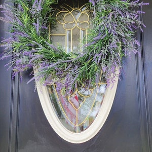 May include: A lavender wreath with green leaves hangs on a black door with a white oval frame and a gold-toned glass window.