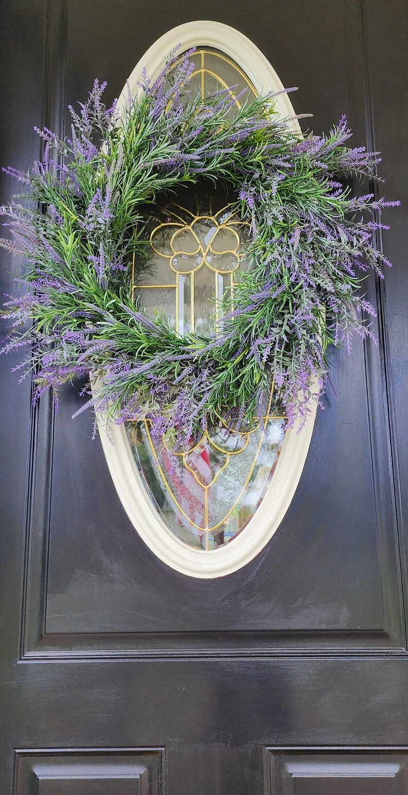 May include: A lavender wreath with green leaves hangs on a black door with a white oval frame and a gold-toned glass window.