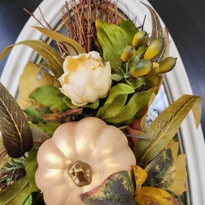 Op de afbeelding: Een gouden pompoen met een witte bloem en groene bladeren in een wit frame. De pompoen is omringd door dennenappels en takken.