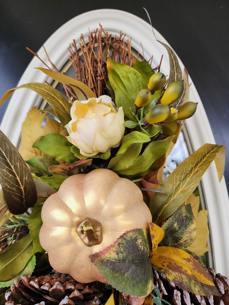 Op de afbeelding: Een gouden pompoen met een steel en bladeren, omringd door herfstbladeren, waaronder dennenappels en een witte bloem.