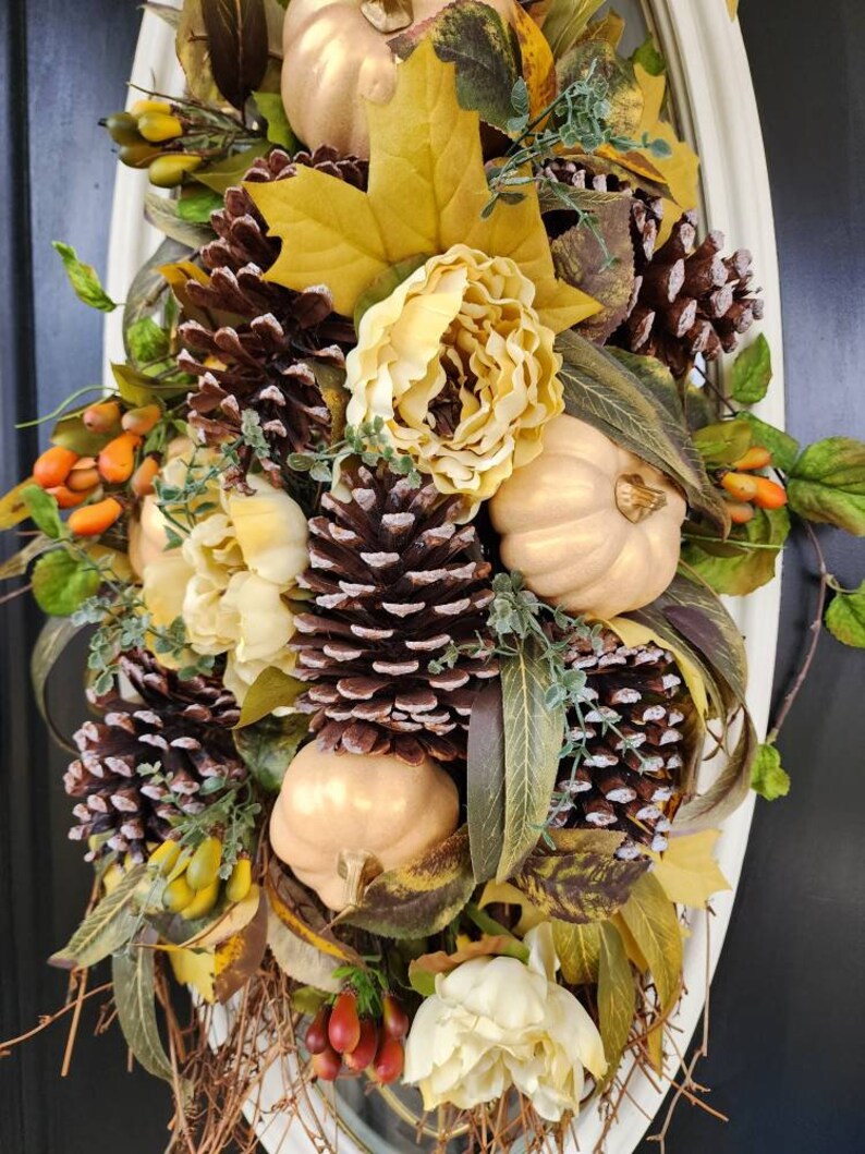 Op de afbeelding: Een herfstkrans met gouden pompoenen, dennenappels en kunstbloemen in cr&egrave;me- en bruintinten. De krans is versierd met herfstbladeren en bessen.