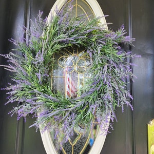 May include: A lavender wreath with green leaves hangs on a black door with a white oval frame. The wreath is made of artificial lavender and green leaves.