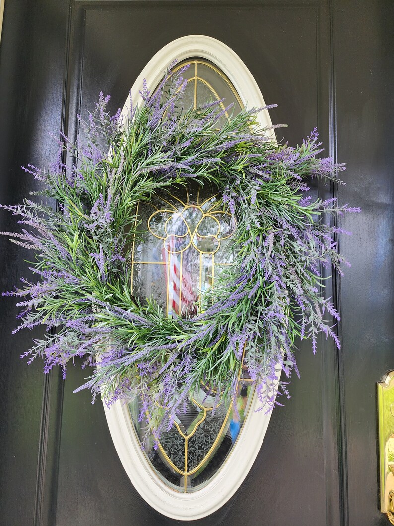 May include: A lavender wreath with green leaves hangs on a black door with a white oval frame. The wreath is made of artificial lavender and green leaves.