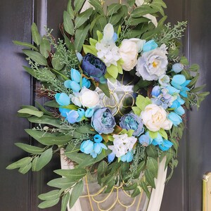 May include: A blue and white floral wreath with green leaves. The wreath is made of artificial flowers and is ideal for a spring or summer door decoration.
