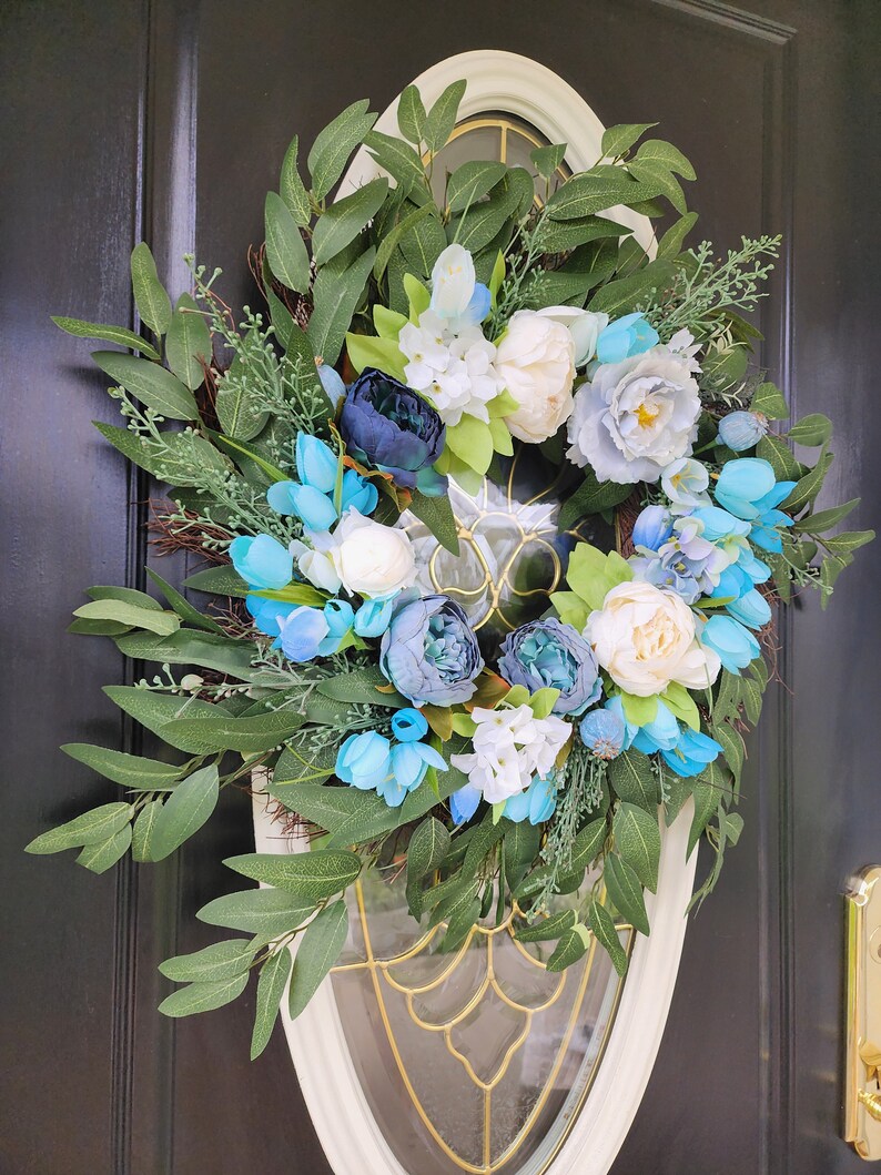 May include: A blue and white floral wreath with green leaves. The wreath is made of artificial flowers and is ideal for a spring or summer door decoration.