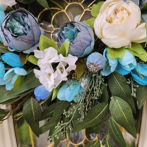 May include: A floral wreath featuring blue, white and green flowers. The wreath is made from artificial flowers and greenery.
