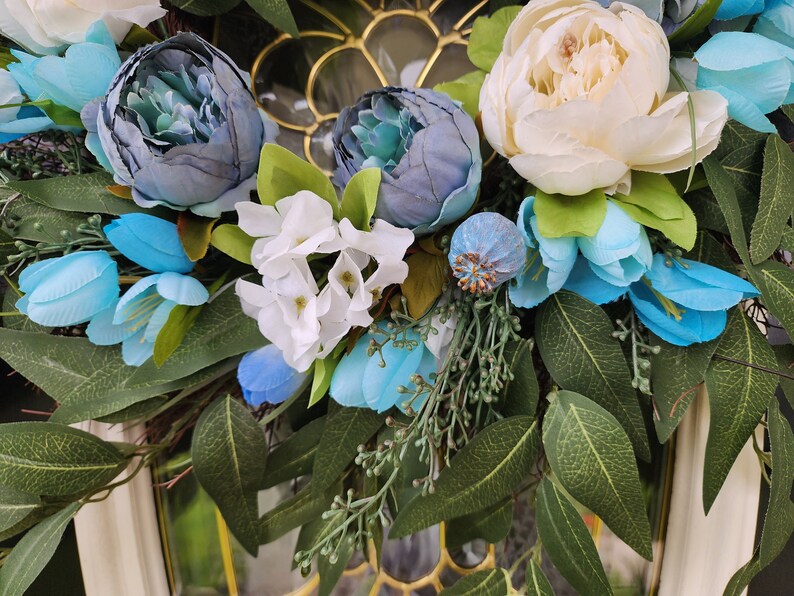 May include: A floral wreath featuring blue, white and green flowers. The wreath is made from artificial flowers and greenery.