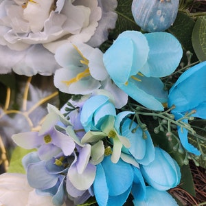 May include: A close-up of a blue and white floral wreath. The wreath is made of artificial flowers, including blue hydrangeas, white peonies, and blue tulips. The wreath is decorated with green leaves and a blue poppy pod.