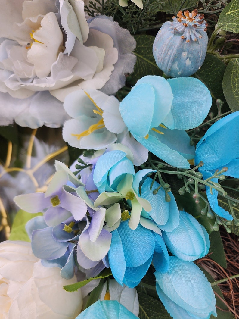 May include: A close-up of a blue and white floral wreath. The wreath is made of artificial flowers, including blue hydrangeas, white peonies, and blue tulips. The wreath is decorated with green leaves and a blue poppy pod.