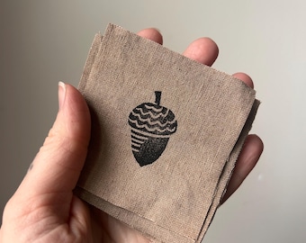 May include: A stack of brown fabric squares with a black acorn design printed on the top square.