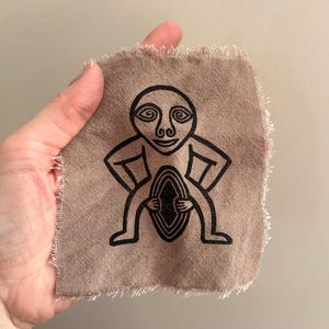 May include: A small, square piece of tan fabric with a black illustration of a stylized human figure. The figure has a large head with spiral eyes, arms akimbo, and a central oval shape. The edges of the fabric are frayed.