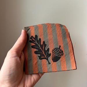 May include: A square piece of fabric featuring vertical stripes in alternating shades of burnt orange and muted olive green. The fabric is printed with a black graphic design depicting a single oak leaf positioned next to a small acorn.
