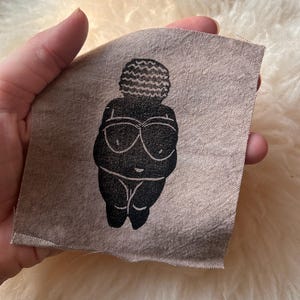 May include: A square piece of light brown, textured fabric featuring a centered black screen-printed graphic of a stylized, rounded female figure with a textured, wavy-patterned hairstyle and prominent circular chest details.