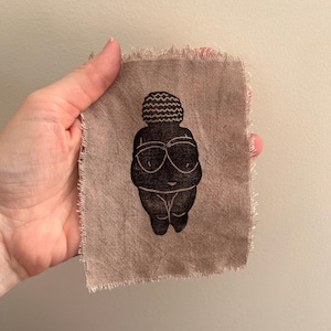 May include: A black print of the Venus of Willendorf on a small, tan fabric square with frayed edges. The image depicts a stylized, faceless female figure with exaggerated features. The fabric is held in a hand against a neutral background.