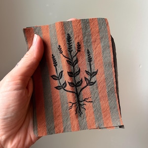 May include: A rectangular piece of fabric featuring vertical stripes in alternating shades of burnt orange and muted olive green. Centered on the fabric is a black ink illustration of a botanical plant with visible roots, leaves, and stems.