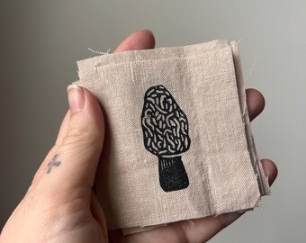 May include: A small, beige linen square with a black and white illustration of a morel mushroom.