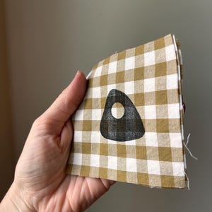 May include: A square piece of fabric featuring a mustard yellow and white gingham check pattern. Centered on the fabric is a black, rounded triangular shape with a small circular cutout in the middle, resembling a planchette design.