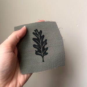 May include: A small, rectangular piece of olive green fabric with a black oak leaf design. The leaf is centered on the fabric, and the edges of the fabric are unfinished. The fabric is held up by a hand.