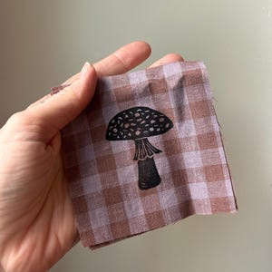 May include: A square piece of fabric featuring a brown and light purple gingham check pattern. Centered on the fabric is a black ink print of a mushroom with a spotted cap and a visible stem, showcasing a detailed botanical illustration style.