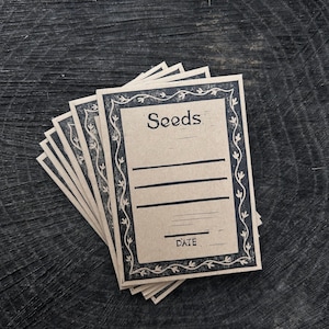 May include: A stack of seed packets with a vintage design. Each packet has the word "Seeds" at the top, with lines for writing and the word "DATE" at the bottom. The packets are beige with a dark blue border.