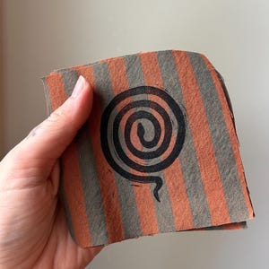 May include: A square fabric coaster featuring vertical stripes in alternating shades of burnt orange and slate gray. Centered on the fabric is a black ink-stamped graphic of a coiled snake forming a spiral shape with a visible tail at the bottom.