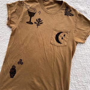 May include: A tan t-shirt with a pocket featuring black graphic designs. The designs include a cocktail glass, a sprig, a moth, a crescent moon with stars, and a stylized figure. The shirt is made of a soft material.