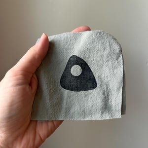 May include: A square piece of light gray linen fabric featuring a centered black triangular shape with a circular cutout in the middle. The fabric has a visible woven texture and raw, slightly uneven edges, displaying a simple geometric print.