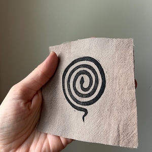 May include: A square piece of light brown, textured fabric featuring a black, hand-printed spiral design that resembles a coiled snake. The fabric has visible woven threads and raw, slightly frayed edges, showcasing a simple, graphic print.