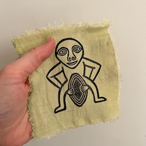May include: A small, light green fabric square with a black graphic print of a stylized human figure. The figure has a large head with spiral eyes and holds a patterned oval shape. The edges of the fabric are frayed.