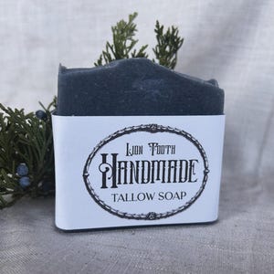 Puede incluir: Una pastilla de jabón de sebo hecha a mano de color gris oscuro con una etiqueta blanca. La etiqueta presenta el texto "Lion Tooth Handmade Tallow Soap" en un marco ovalado decorativo. Ramas verdes y bayas en el fondo.
