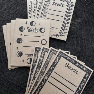 Op de afbeelding: Een verzameling zaadzakjes van kraftpapier met zwarte inktontwerpen. Sommige zakjes tonen maanfasen, andere bloemenranden. Elk zakje is gemarkeerd met "Seeds" en bevat lijnen voor details en een "Datum" sectie.