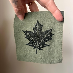May include: A square, olive green fabric patch featuring a black maple leaf design. The leaf is centered on the fabric, with detailed veins and a classic shape. The fabric has a textured appearance.