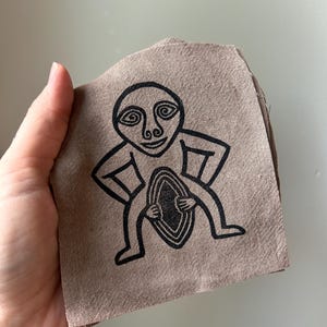 May include: A square piece of light brown, textured fabric featuring a black line-art illustration of a stylized humanoid figure with spiral eyes, hands on hips, and holding an oval shape in the center of its torso with both hands.
