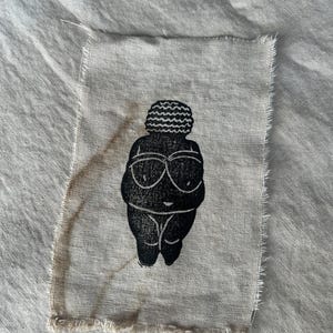 May include: A black and white illustration of a person with a wavy hairstyle, wearing sunglasses, printed on a beige fabric patch.