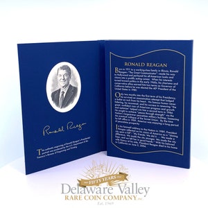 May include: A blue commemorative folder featuring a black and white portrait of Ronald Reagan in an oval frame, his gold signature, and a biography printed in gold text. The folder includes text about his life, presidency, and death in 2004.